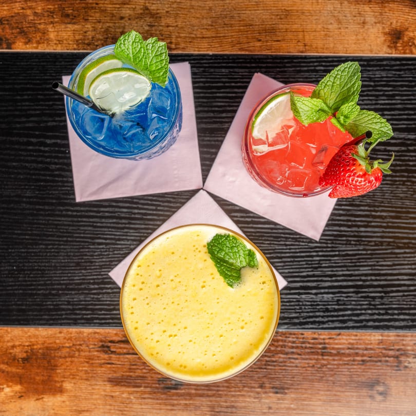 Three colorful cocktails, blue, red, and yellow, garnished with mint, lime, and strawberry on a wooden table.