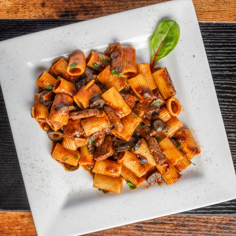 Rigatoni pasta with beef, black olives, and red sauce, garnished with a single green leaf.