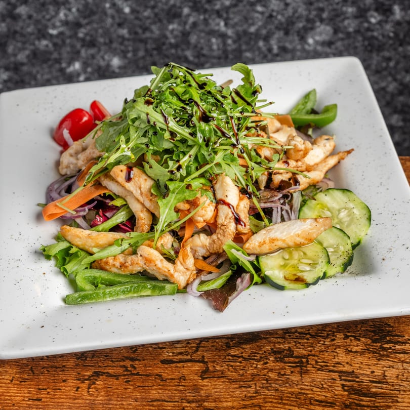 Pastaloni's chicken salad with arugula, cherry tomatoes, cucumbers, and balsamic glaze on a white plate.