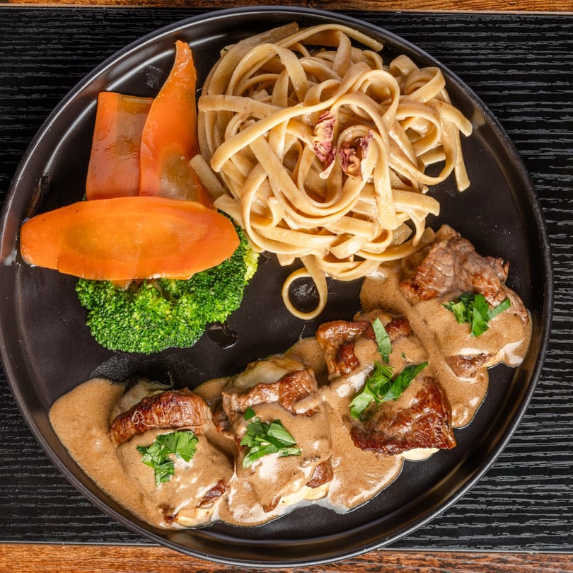 Pastaloni's Italian dish with tagliatelle, meat in creamy sauce, broccoli, and carrots on a black plate.