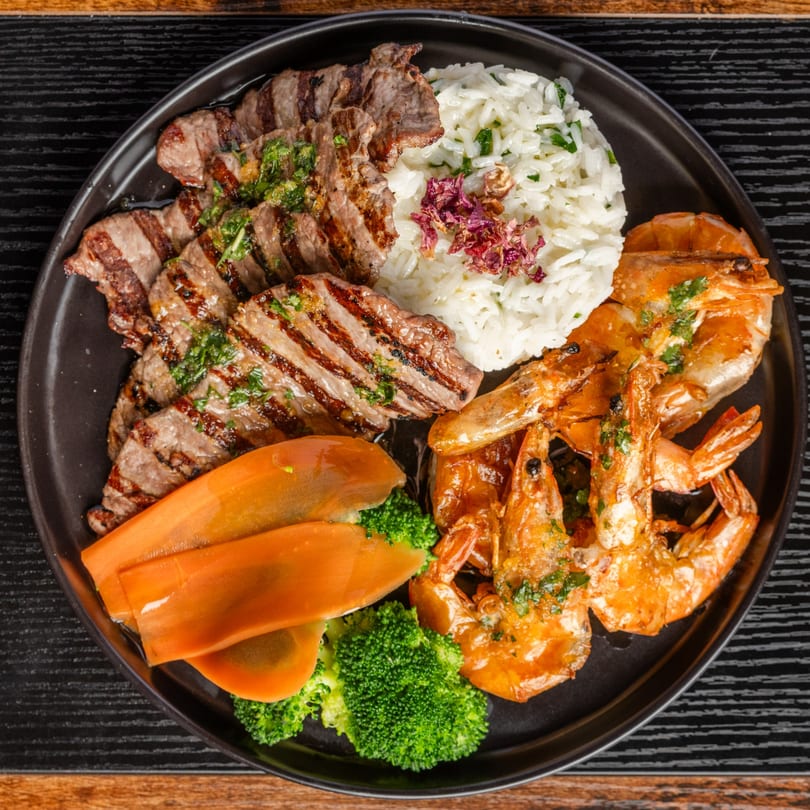 Grilled steak, shrimp, white rice, carrots, and broccoli on a black plate.